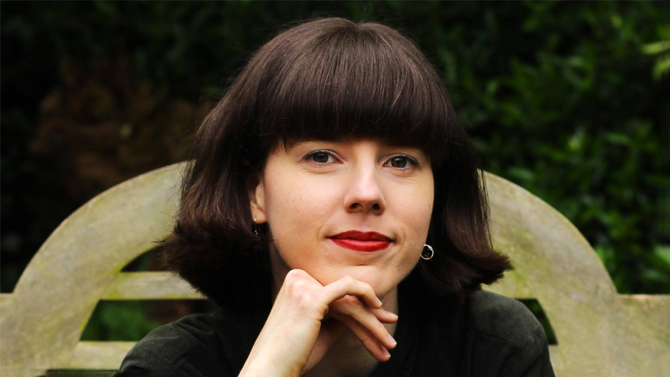 A women with short dark hair, learning on her hand, sitting outside, smiling at the camera.